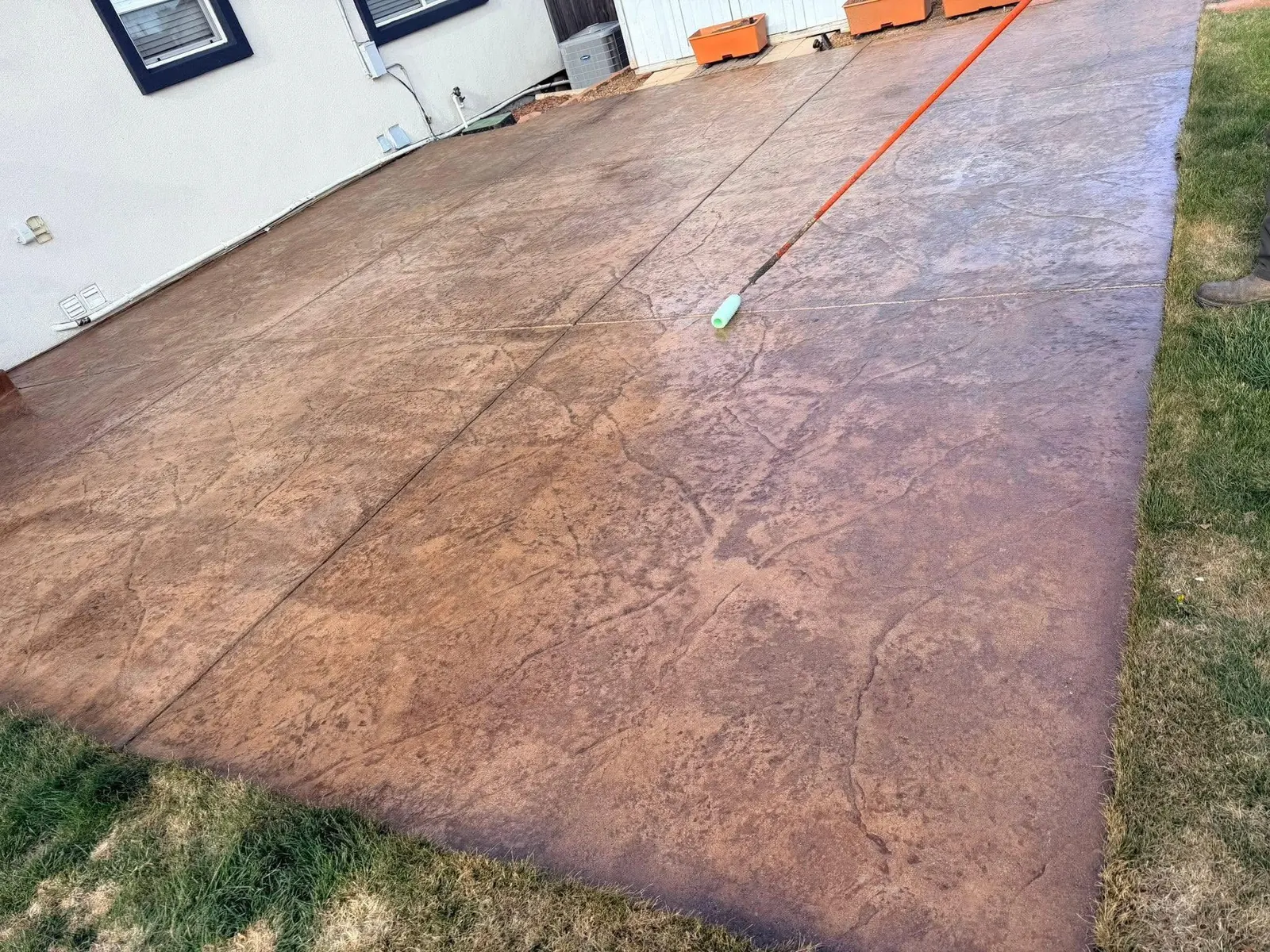 A large russet stamped concrete patio being rolled with sealer, the wet half noticeably darker than the dry half.