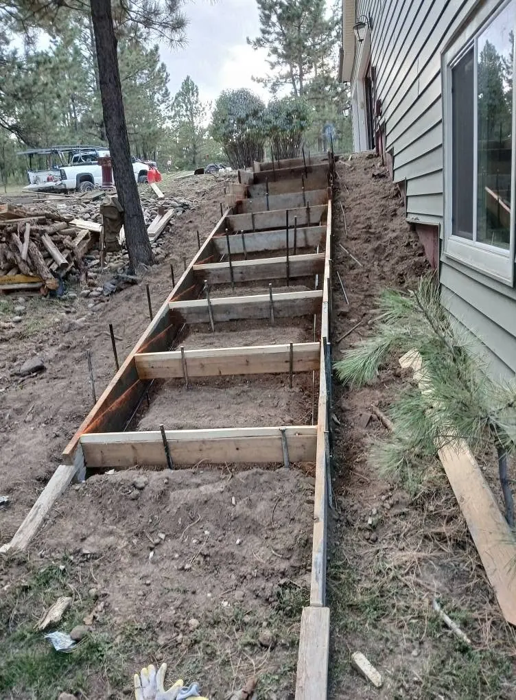 A flight of steps formed in timber with steel rebar standing up through it, cut into a dirt slope beside a house in the pines.