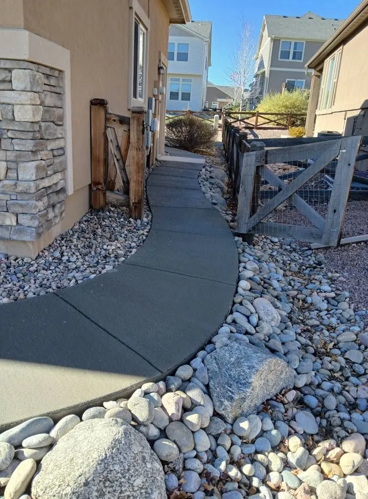 The same side walkway finished — a smooth curving concrete path sweeping between the house and the river rock.