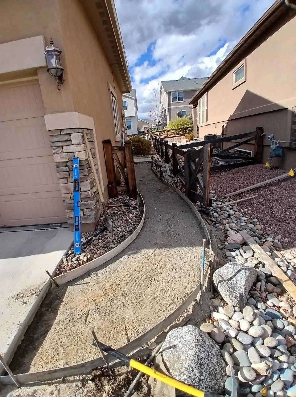 A curved side walkway formed and ready to pour, with compacted base between a stone-veneer wall and a river-rock bed.