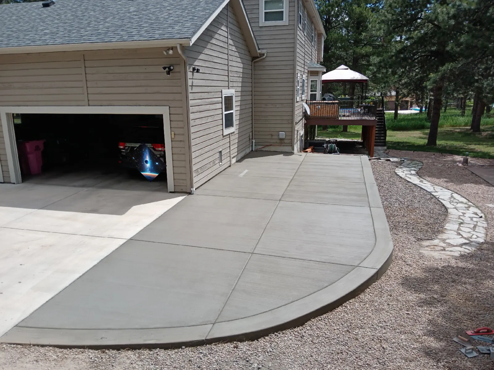 A broom-finished concrete driveway extension beside a two-car garage, ending in a smooth curve against a gravel path.
